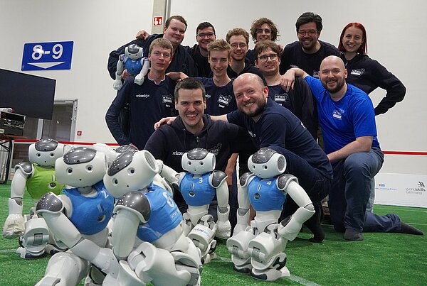 Die HTWK Robots im Wettkampfjahr 2025. Gruppe junger Leute mit humanoiden Robotern im Vordergrund