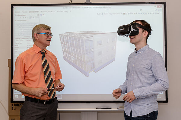 Professor Ulrich Möller und Nachwuchsforscher Tom Radisch vor einem virtuellen Modell eines Gebäudes. Radisch trägt eine Virtual-Reality-Brille.