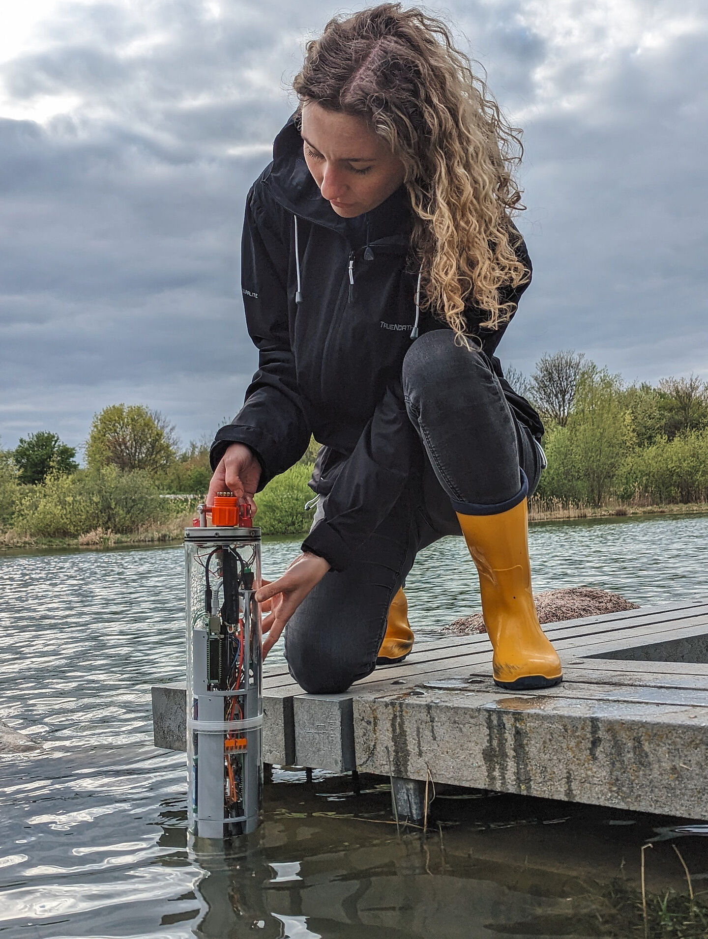 eine Frau hockt am Wasser und hält eine Messsonde in einen See