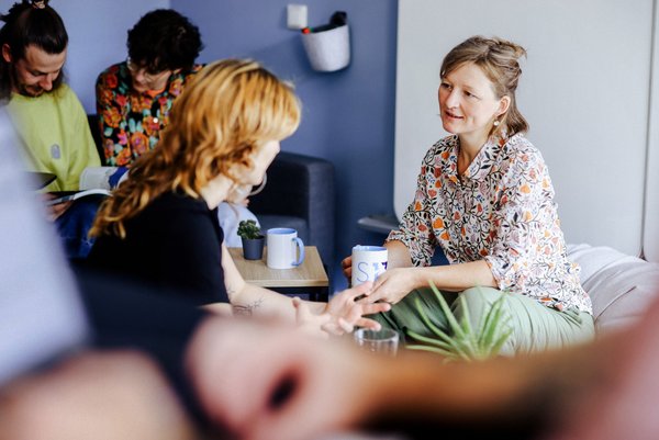 Mentoring Mehrere Personen sitzen in einem Besprechungsrauum. Zwei Frauen im Vordergrund unterhalten sich, im Hintergrund lesen zwei Personen ein Buch.