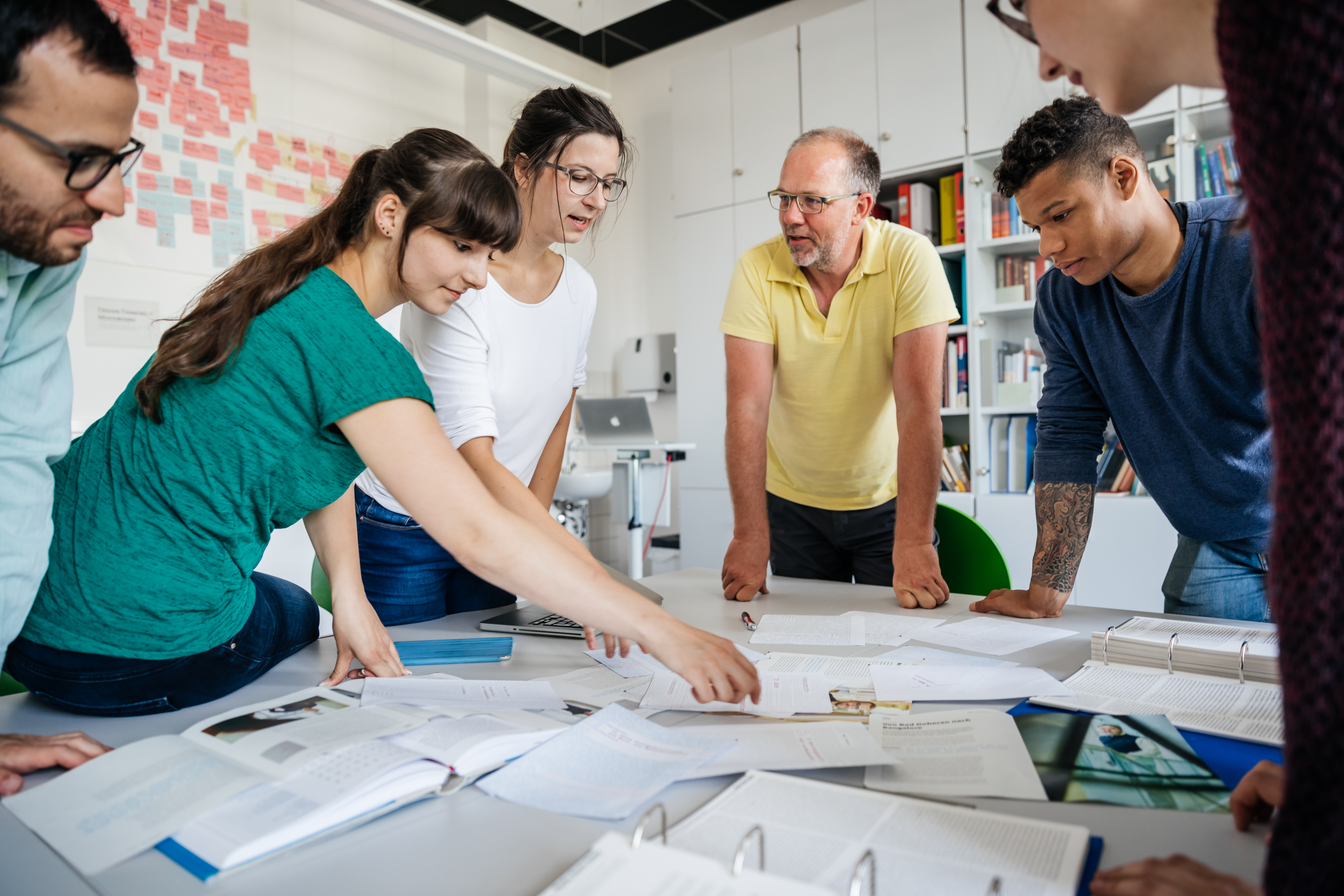 gemeinschaftliche Arbeit Sechs Personen stehen um einen Tisch mit vielen offenen Unterlagen und diskutieren