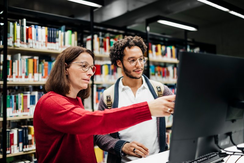 Eine Frau zeigt einem Mann in einer Bibliothek etwas auf einem Computer.